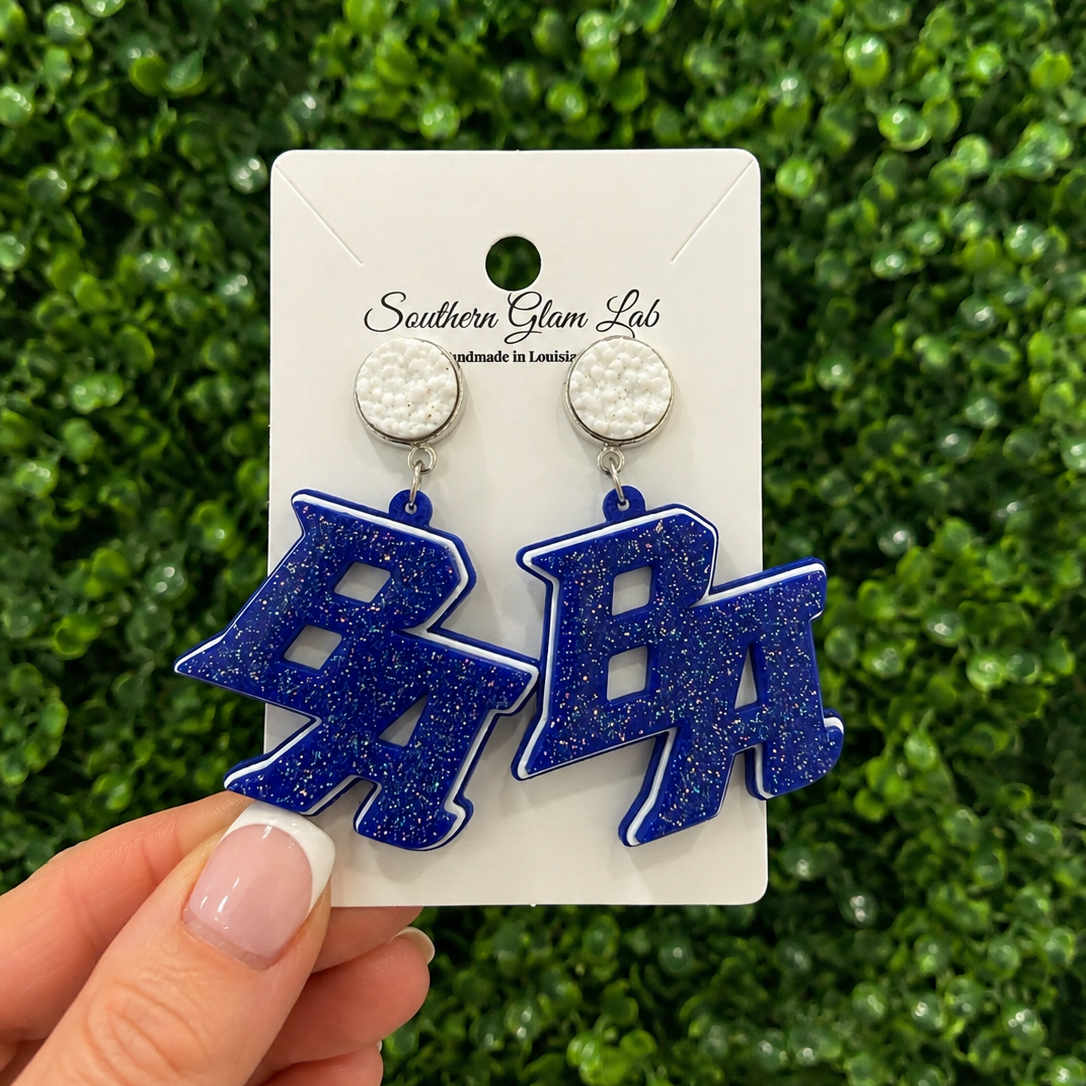 A hand holds a white display card featuring a pair of blue glitter letter earrings shaped like the letters 'BA' against a green foliage background. The card is branded 'Southern Glam Lab'.