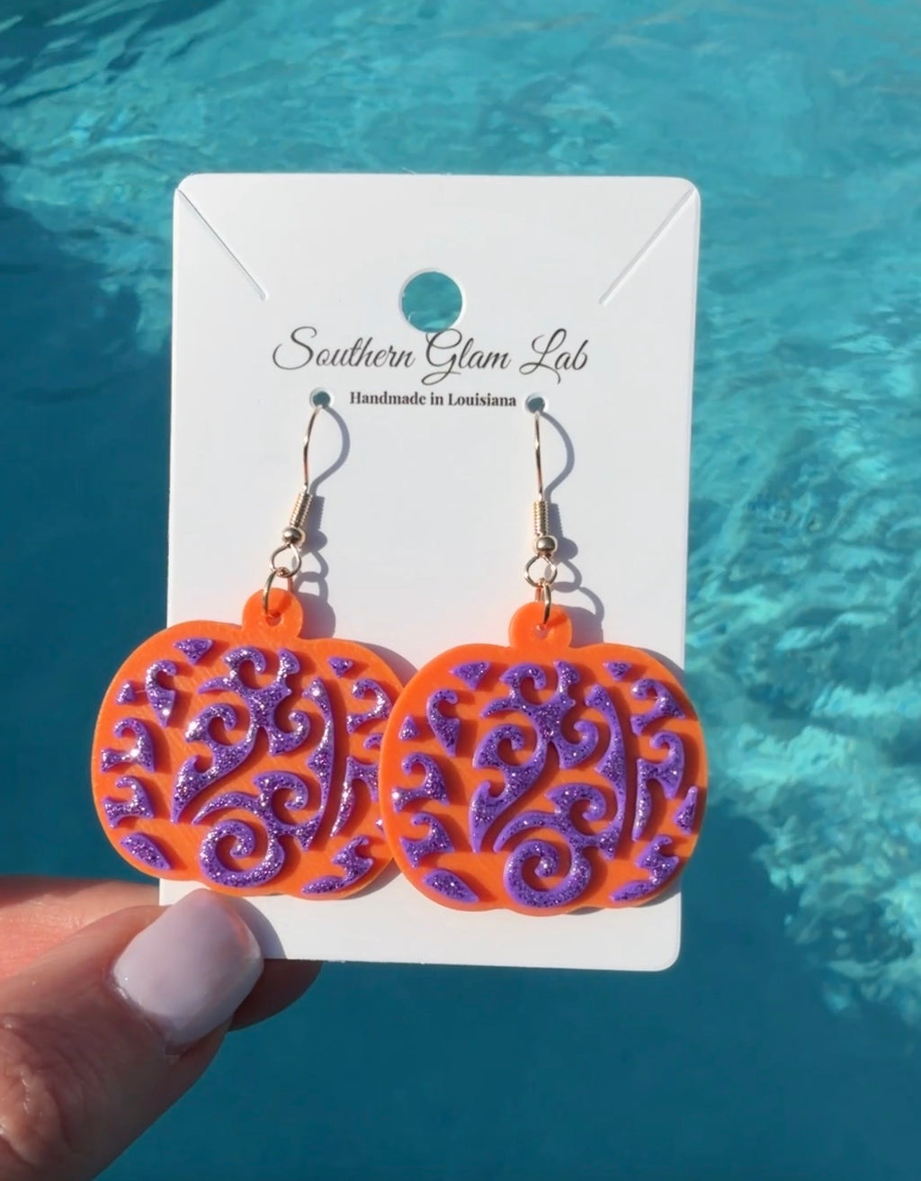 A pair of dangling earrings displayed on a white card held by a hand, with a bright blue swimming-pool background. The earrings have glittery orange and purple pumpkin-shaped charms with swirling cutout patterns, attached to gold hooks.