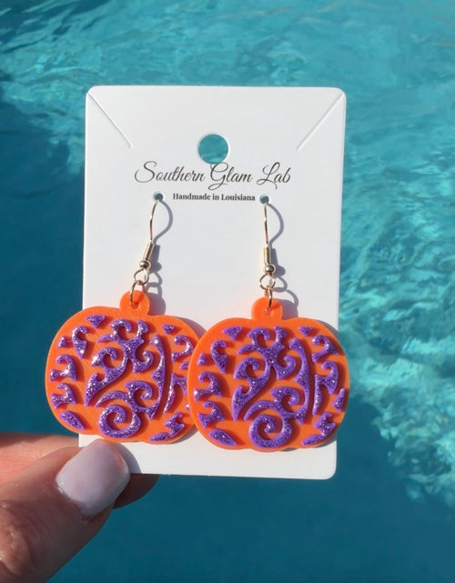 A pair of dangling earrings displayed on a white card held by a hand, with a bright blue swimming-pool background. The earrings have glittery orange and purple pumpkin-shaped charms with swirling cutout patterns, attached to gold hooks.
