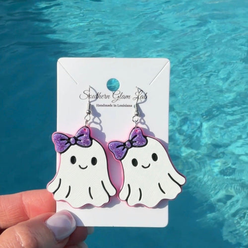 A hand holds a pair of white ghost-shaped dangle earrings with purple bows on a white display card, set against a bright blue swimming pool background.