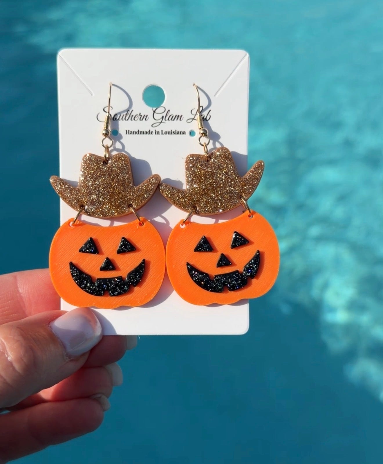 A pair of Halloween drop earrings featuring orange jack-o'-lantern shapes with black glitter facial details and glittery gold cowboy hats, displayed on a white card held by a hand against a blue pool background.