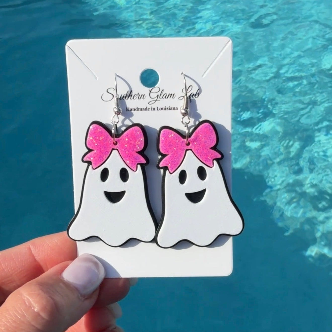 A hand holds a pair of ghost-shaped dangle earrings with pink glitter bows on a white display card, with a bright blue swimming pool in the background.