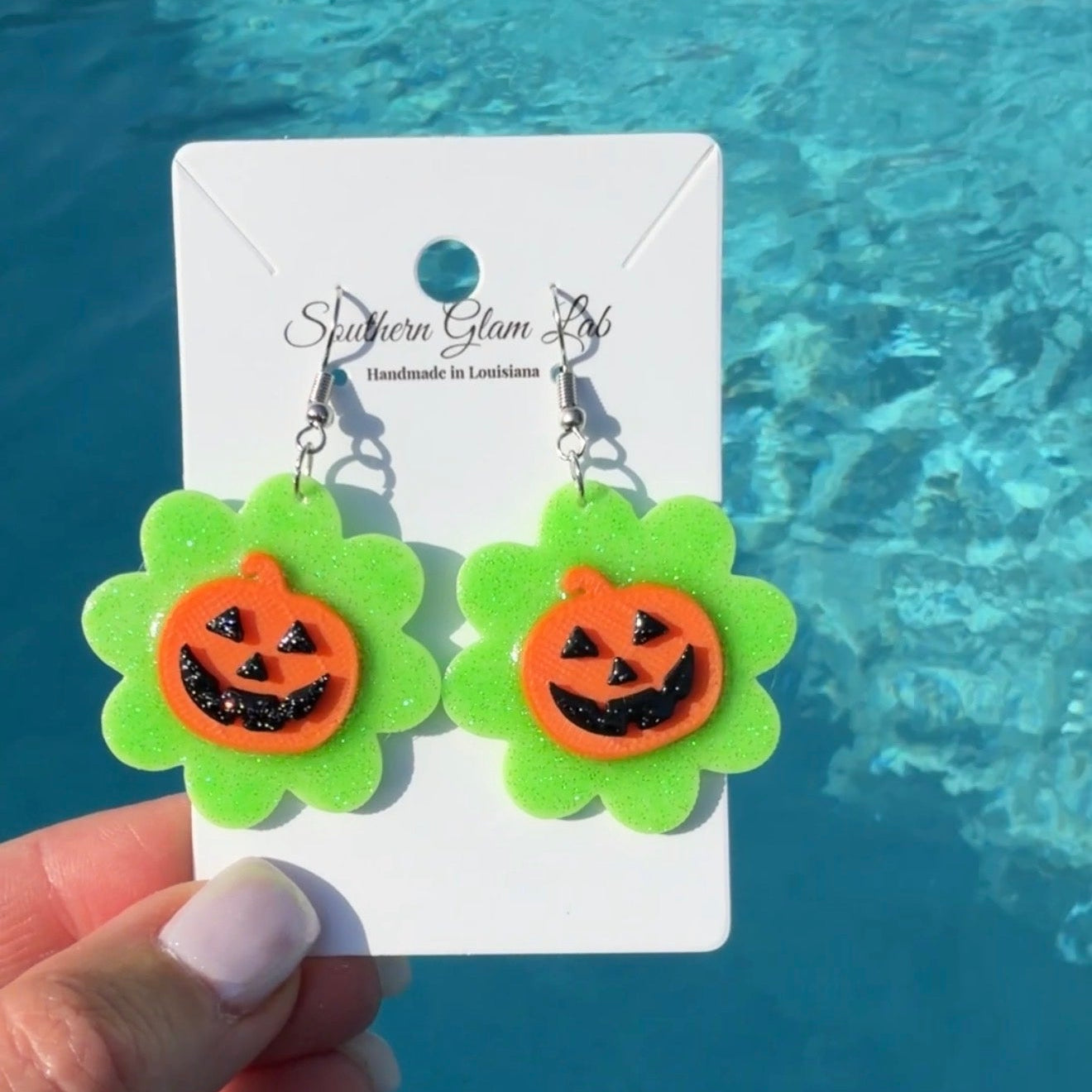 A pair of Halloween dangle earrings held by a hand in front of a swimming pool. The earrings feature bright green scalloped flower-shaped drops with orange pumpkin faces and black details, hanging from silver hooks on a white display card labeled Southern Glam Lab.
