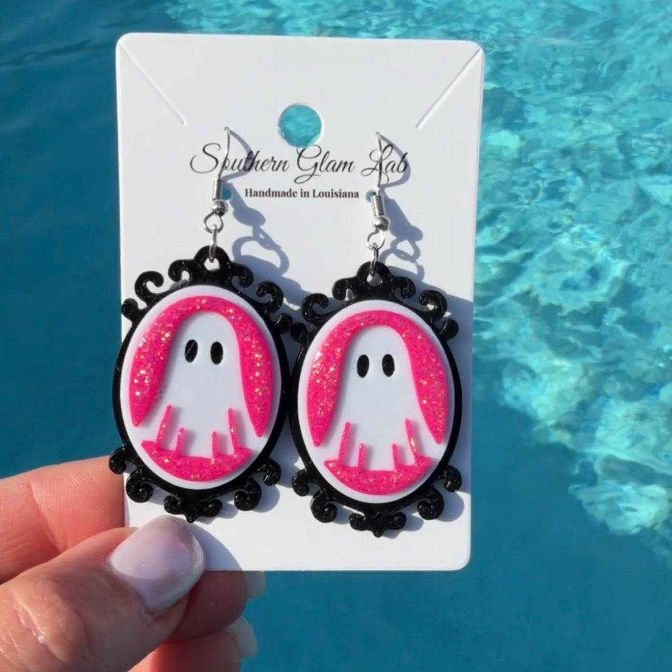 A hand holds a pair of dangling earrings on a white display card in front of bright blue water. The earrings have black scalloped oval frames with pink glitter-filled centers featuring white ghost cutouts with black eyes.