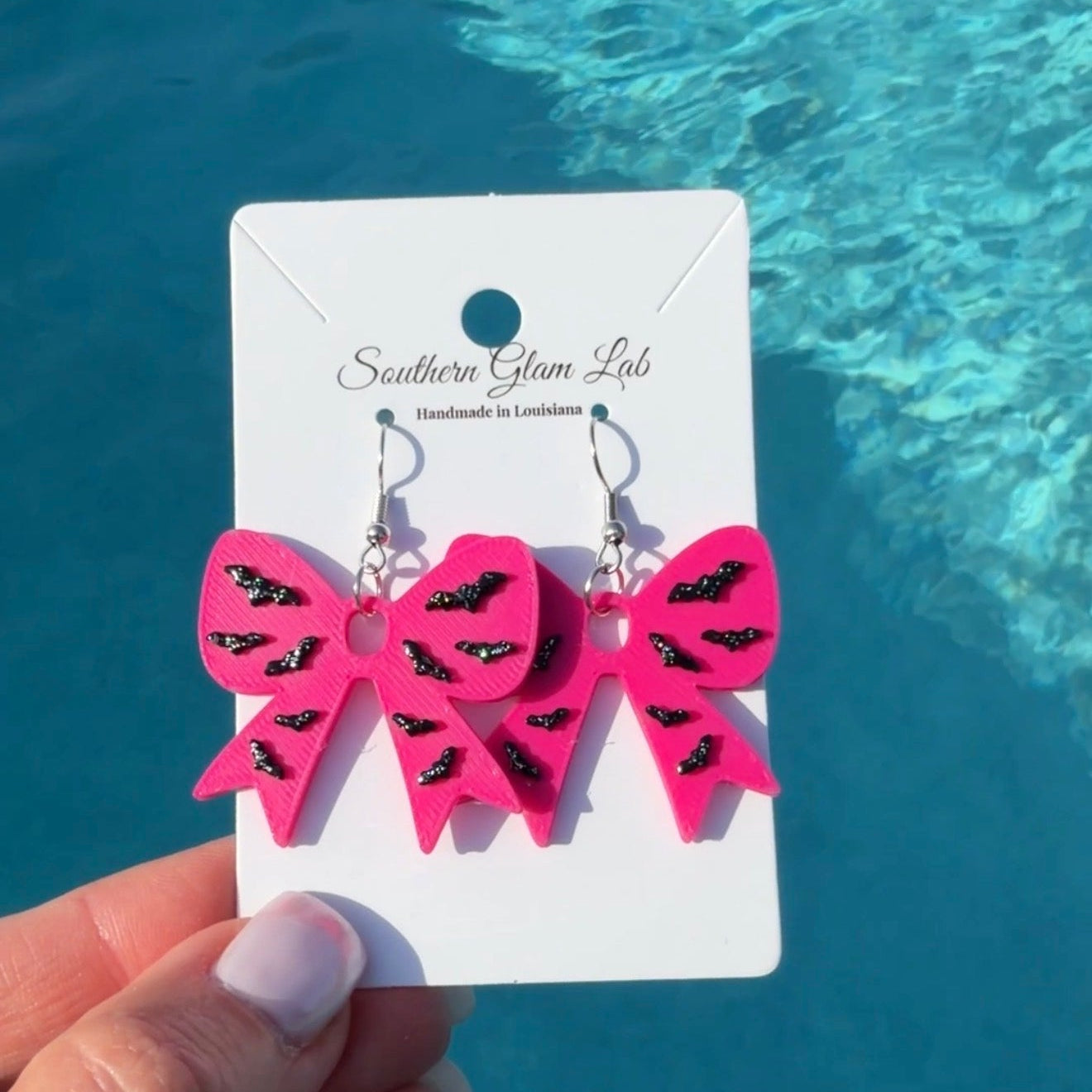 Bright pink bow-shaped dangle earrings with black glitter accents, displayed on a white card labeled Southern Glam Lab and held by a hand in front of a swimming pool background.