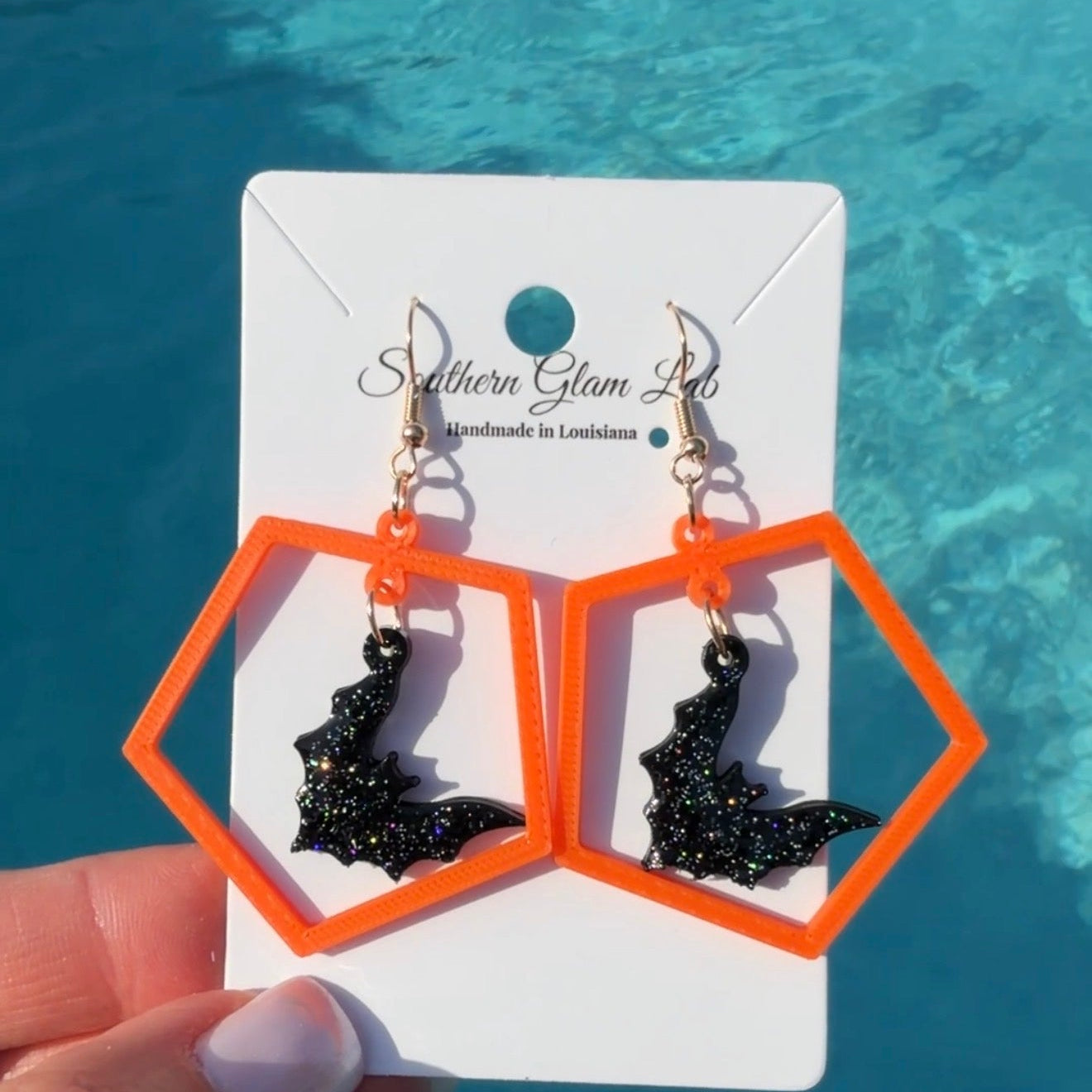A pair of Halloween dangle earrings with black glitter bat charms inside bright orange hexagonal frames, displayed on a white card held by a hand against a swimming pool background.
