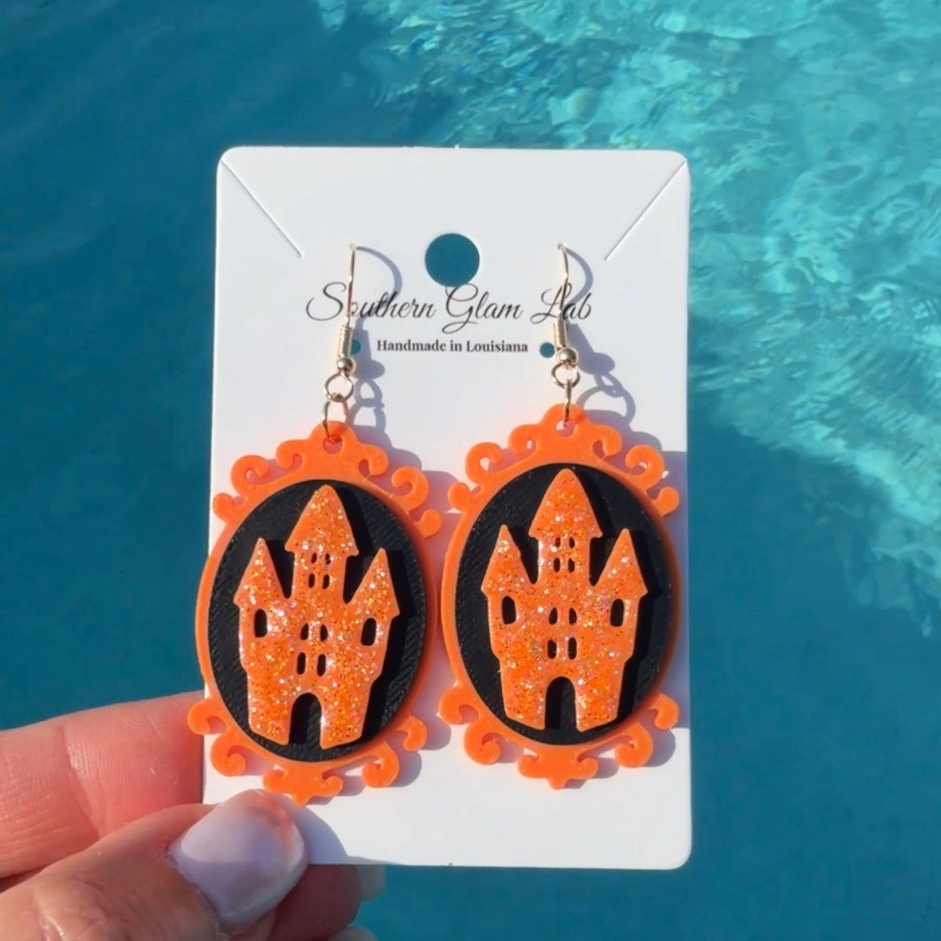 A pair of orange and black Halloween dangle earrings with castle motifs, displayed on a white card held by a hand against a bright blue swimming pool background.