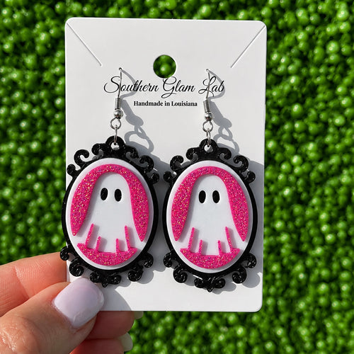 A pair of gothic-style dangle earrings with pink glitter ghost charms inside black oval frames, displayed on a white card held by a hand against a green textured background. The card is branded 'Southern Glam Lab'.
