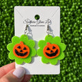 A pair of Halloween dangle earrings held by a hand against a green grass-like background. Each earring has an orange pumpkin face with black glitter details on a bright green flower-shaped backing, attached to silver hooks and displayed on a white card labeled Southern Glam Labs.