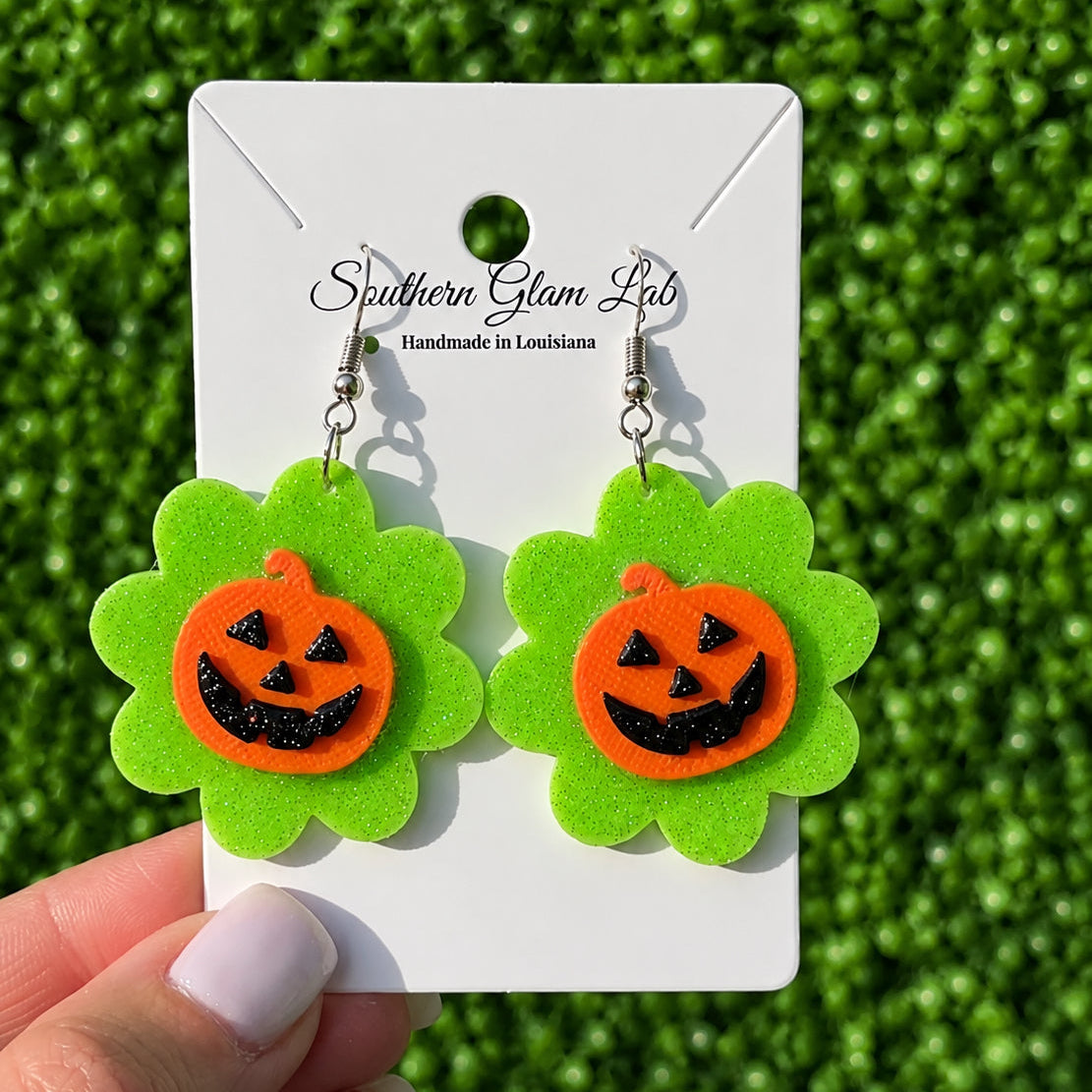 A pair of Halloween dangle earrings held by a hand against a green grass-like background. Each earring has an orange pumpkin face with black glitter details on a bright green flower-shaped backing, attached to silver hooks and displayed on a white card labeled Southern Glam Labs.