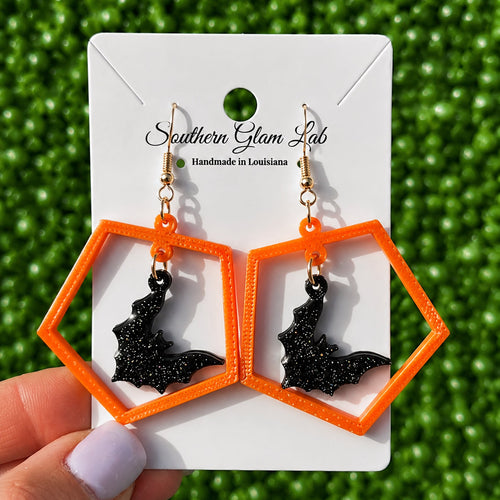 A pair of orange and black 3D-printed geometric dangle earrings with bat charms, displayed on a white card held by a hand against a green textured background. The card is branded 'Southern Glam Lab'.