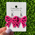 A pair of bright pink bow-shaped dangle earrings with black glitter accents, displayed on a white card labeled Southern Glam Lab, held by a hand against a green grassy background.