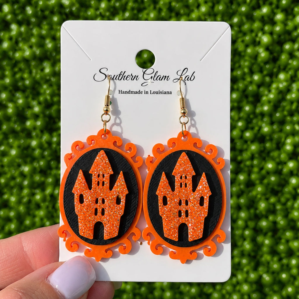 A pair of orange and black Halloween-themed dangle earrings featuring a glittery orange haunted castle on a black oval background, displayed on a white card held by a hand against a green textured backdrop.