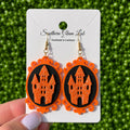 A pair of orange and black Halloween-themed dangle earrings featuring a glittery orange haunted castle on a black oval background, displayed on a white card held by a hand against a green textured backdrop.