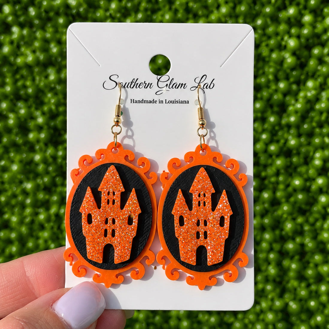A pair of orange and black Halloween-themed dangle earrings featuring a glittery orange haunted castle on a black oval background, displayed on a white card held by a hand against a green textured backdrop.