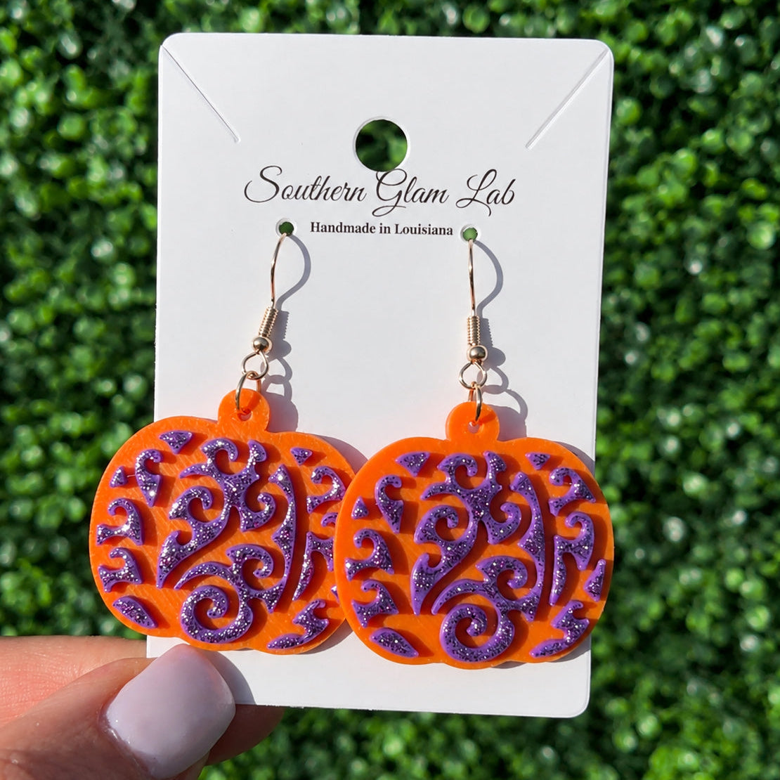 A pair of orange and purple glitter dangle earrings displayed on a white card labeled 'Southern Glam Lab,' held by a hand against a green hedge background.