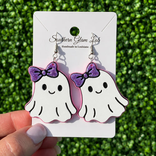 A pair of white ghost-shaped dangle earrings with purple glitter bows, displayed on a white card labeled Southern Glam Lab, held by a hand against a green grass background.