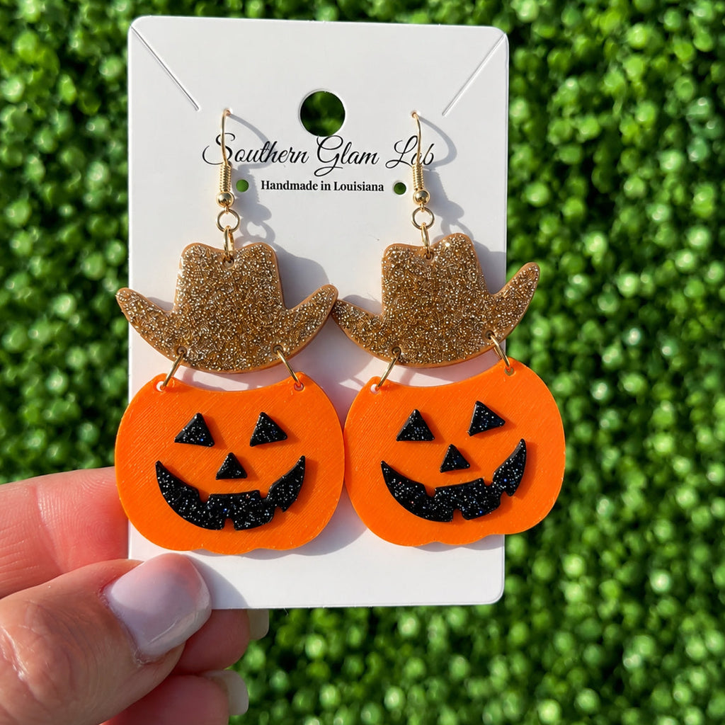 A pair of Halloween-themed dangling earrings displayed on a white card held by a hand. Each earring has a glittery gold cowboy hat above an orange pumpkin face with black glitter details.
