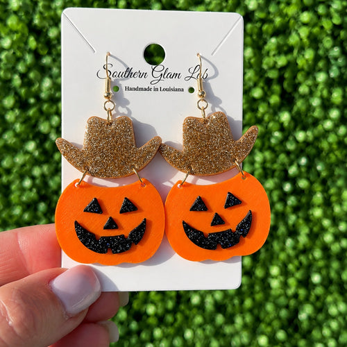 A pair of Halloween-themed dangling earrings displayed on a white card held by a hand. Each earring has a glittery gold cowboy hat above an orange pumpkin face with black glitter details.