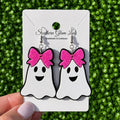 A pair of ghost-shaped dangle earrings with bright pink glitter bows, displayed on a white card held by a hand against a green textured background. The card is branded 'Southern Glam Lulz'.