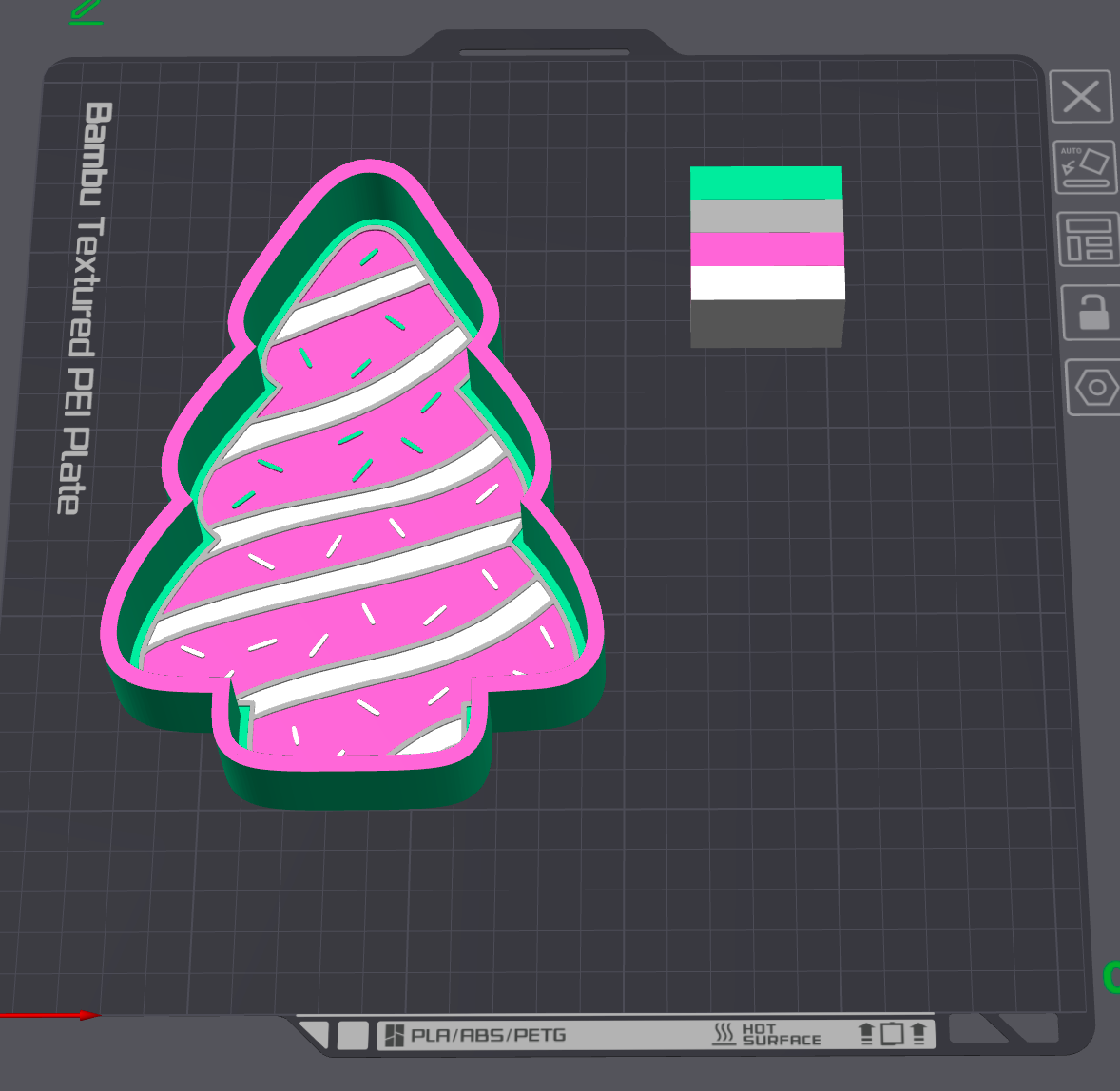 A 3D digital preview of a textured PET plate shaped like a Christmas tree, featuring pink frosting-like stripes, white sprinkles, a green inner layer, and a dark gray outer frame. It is displayed on a dark gray grid design screen with a color swatch in the top right.